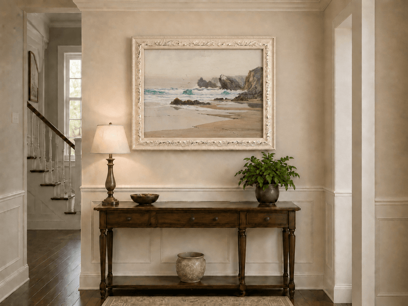 Framed hand-painted coastal beach oil painting with crashing waves and cliffside rocks displayed above an entryway console table.