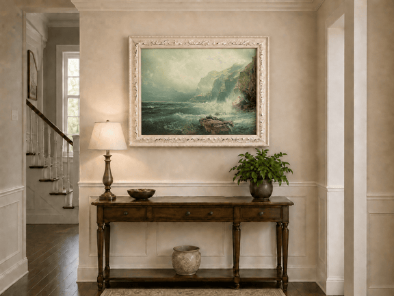 Framed hand-painted coastal cliff oil painting with crashing waves beneath a misty sky displayed above an entryway console table.