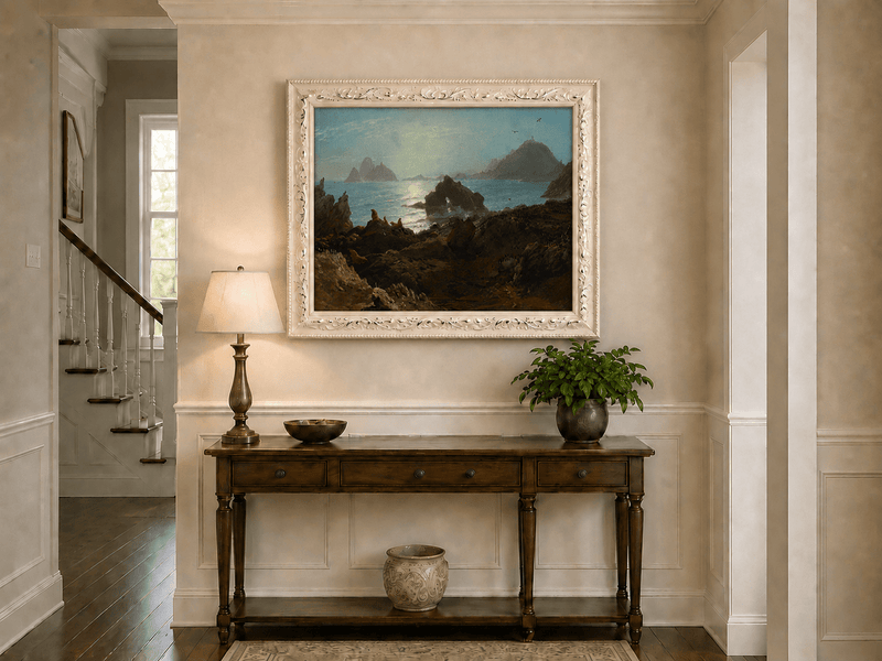 Framed hand-painted coastal oil painting with a rocky shoreline, wildlife and distant sea stacks displayed above an entryway console table.
