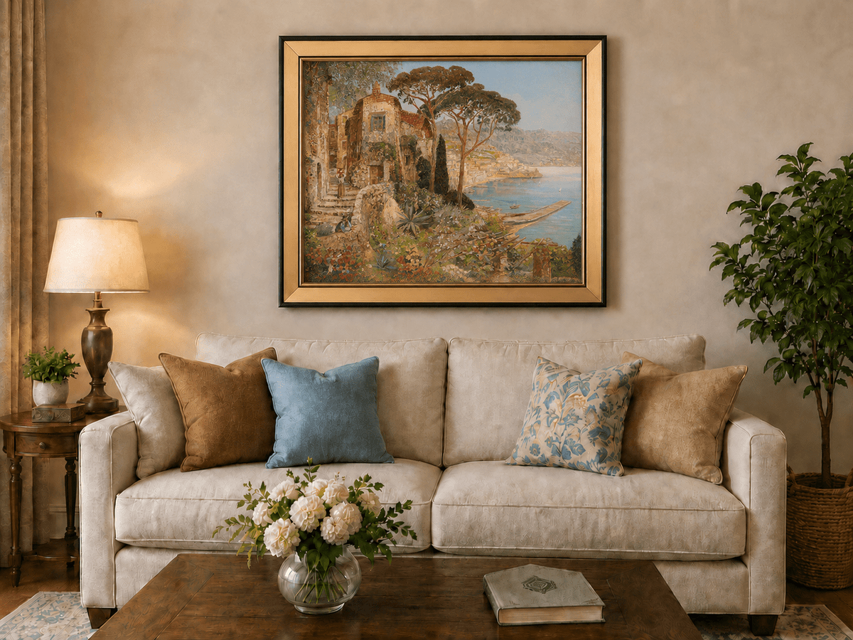 Framed hand-painted coastal village oil painting with flowers and blue sea displayed above a neutral living room sofa.