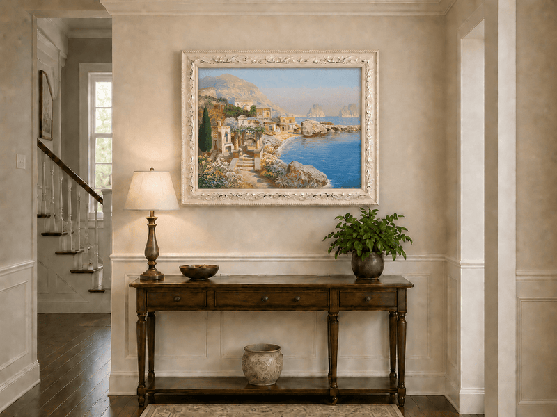 Framed hand-painted coastal village oil painting with blue sea and a rocky shoreline displayed above an entryway console table.