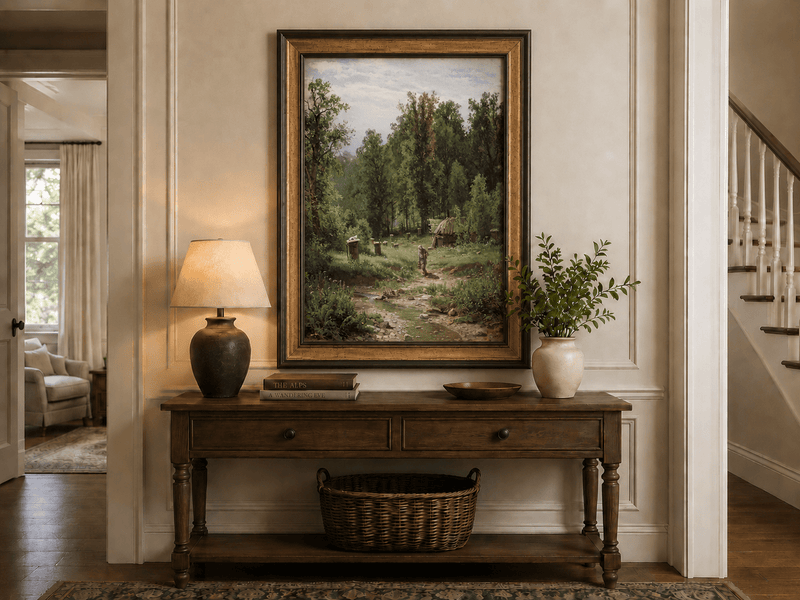 Framed hand-painted forest cottage oil painting with a stream, path and walking figure displayed above an entryway console table.
