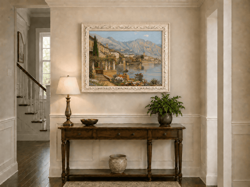 Framed hand-painted lakeside village oil painting with flowers and mountains displayed above an entryway console table.