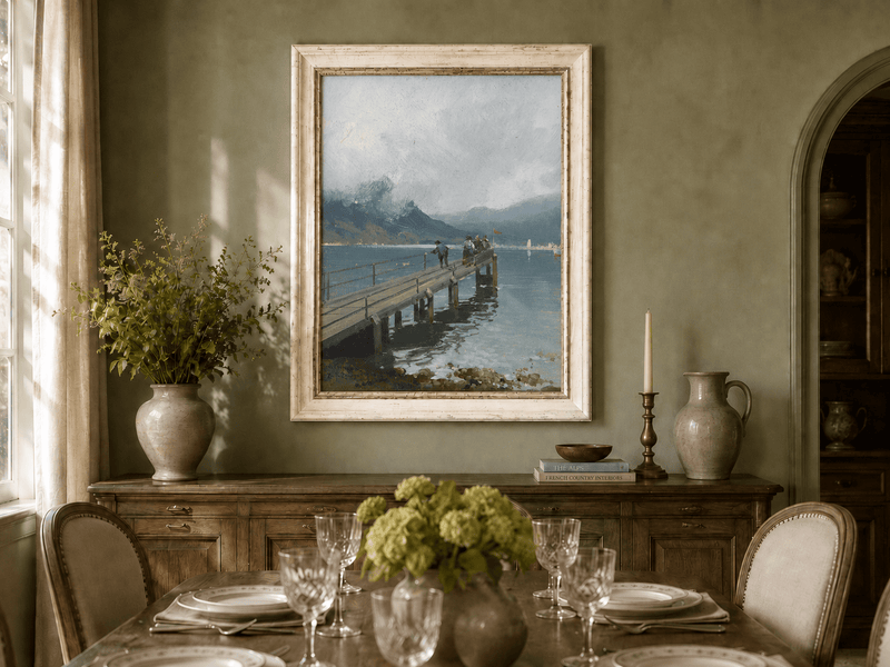 Alpine lake pier with figures and boats oil landscape artwork shown on a wall in a styled home interior.