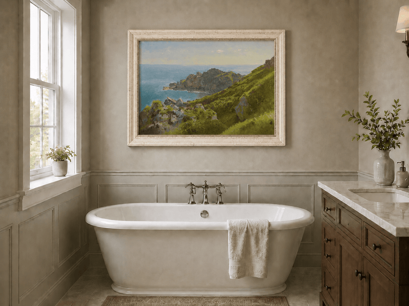 Framed hand-painted coastal landscape oil painting with green hills, rocky headland and ocean displayed above a freestanding bathtub.