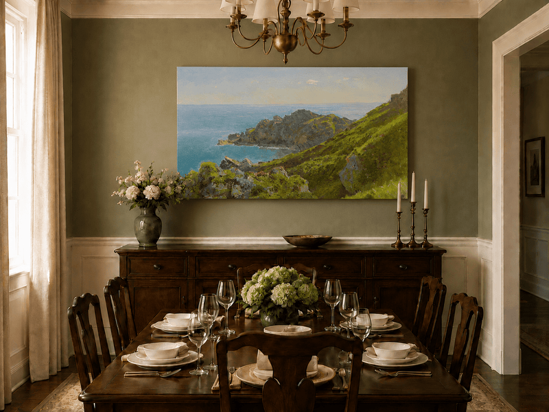 Unframed hand-painted coastal landscape oil painting with green hills, rocky headland and ocean displayed above a formal dining table.