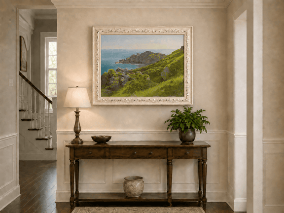 Framed hand-painted coastal landscape oil painting with green hills, rocky headland and ocean displayed above an entryway console table.