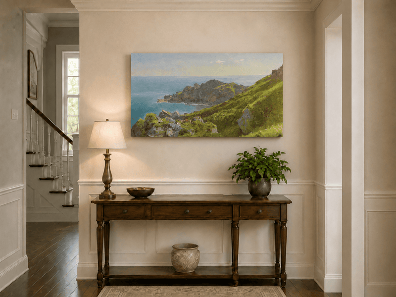 Unframed hand-painted coastal landscape oil painting with green hills, rocky headland and ocean displayed above an entryway console table.