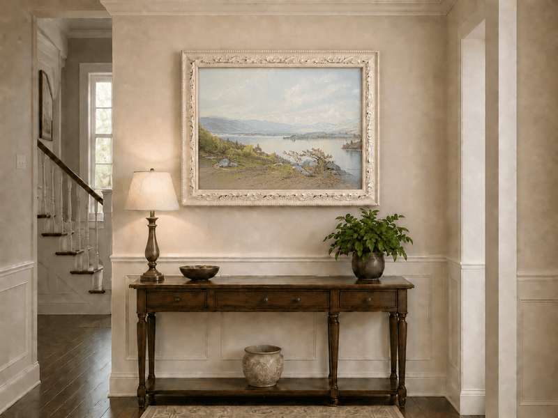 Framed hand-painted expansive lake oil painting with distant mountains, trees and calm water displayed above an entryway console table.