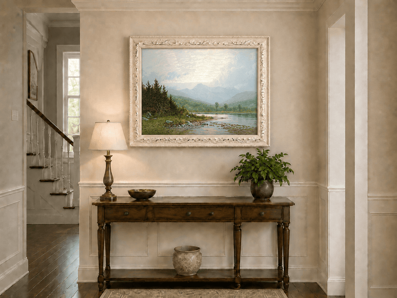 Framed hand-painted lake shore oil painting with pine shade, still water and mountain peaks displayed above an entryway console table.
