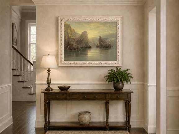 Framed hand-painted seascape oil painting with rock cliffs, calm water and glowing skies displayed above an entryway console table.