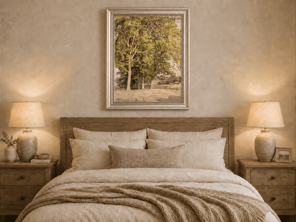 Framed hand-painted rural landscape oil painting with leafy trees and a grassy field displayed above a wooden bed in a neutral bedroom.
