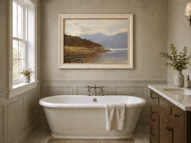 Framed hand-painted lake shore oil painting with rocky banks, vegetation and mountain ranges displayed above a freestanding bathtub.