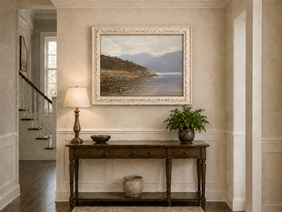 Framed hand-painted lake shore oil painting with rocky banks, vegetation and mountain ranges displayed above an entryway console table.