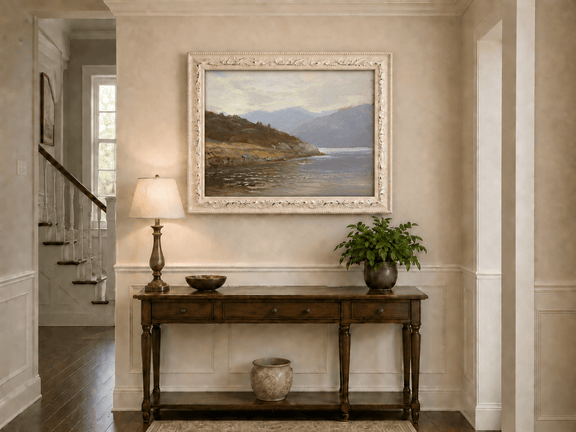 Framed hand-painted lake shore oil painting with rocky banks, vegetation and mountain ranges displayed above an entryway console table.