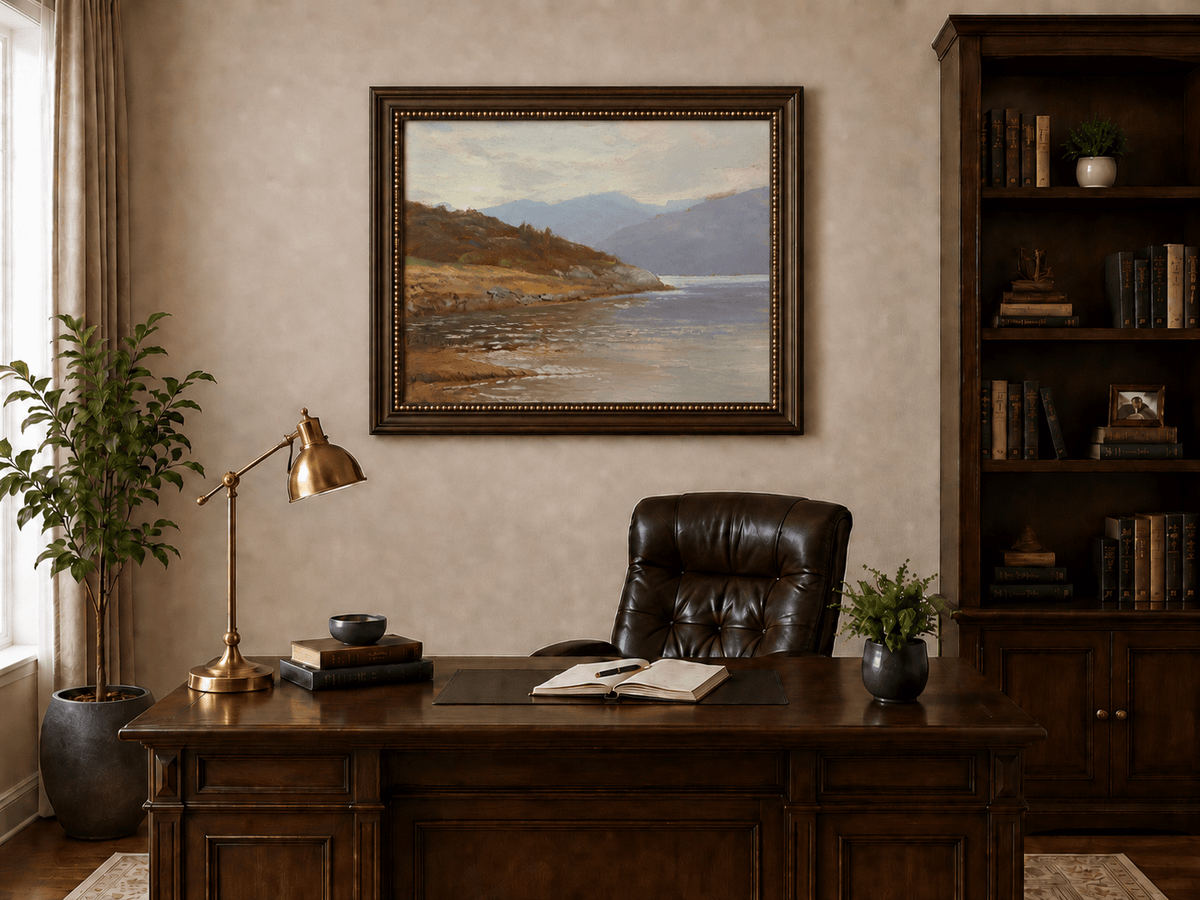 Framed hand-painted lake shore oil painting with rocky banks, vegetation and mountain ranges displayed above a traditional home office desk.