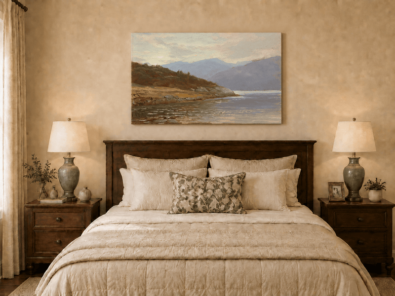 Unframed hand-painted lake shore oil painting with rocky banks, vegetation and mountain ranges displayed above a wooden bed in a neutral bedroom.