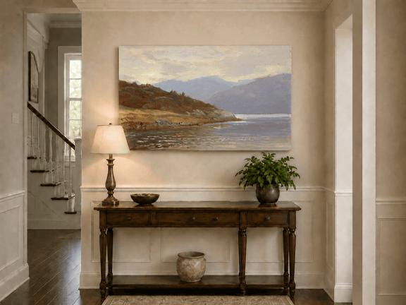 Unframed hand-painted lake shore oil painting with rocky banks, vegetation and mountain ranges displayed above an entryway console table.