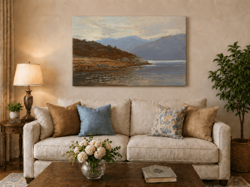 Unframed hand-painted lake shore oil painting with rocky banks, vegetation and mountain ranges displayed above a neutral living room sofa.