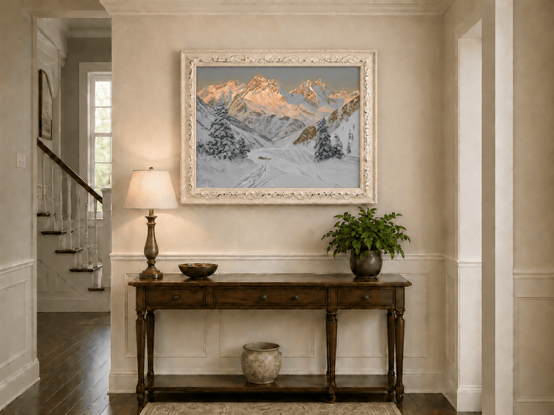 Framed hand-painted alpine winter oil painting with a snowy hut, winding tracks and sunlit peaks displayed above an entryway console table.
