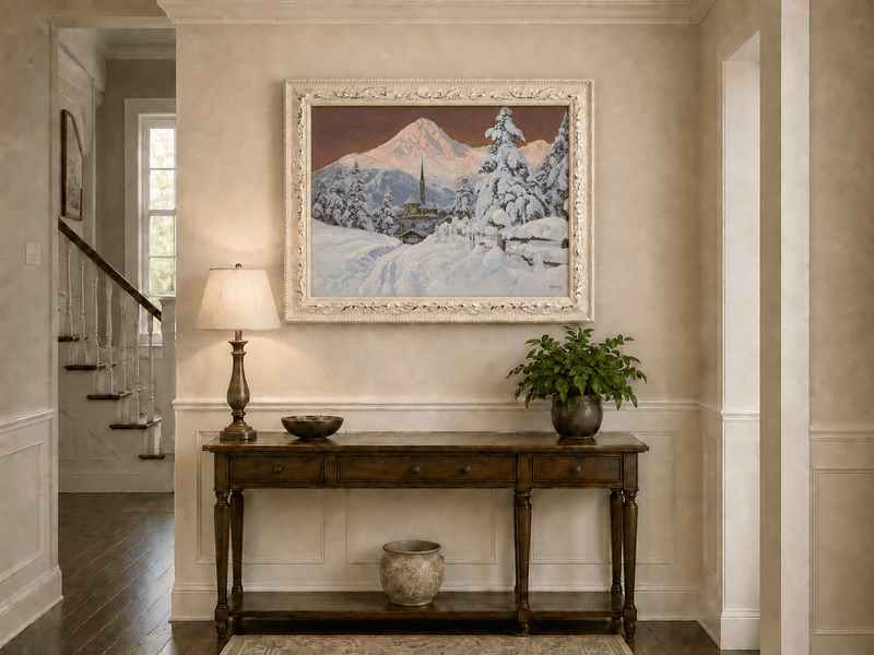 Framed hand-painted snowy alpine village oil painting with a church and pink mountain peaks displayed above an entryway console table.
