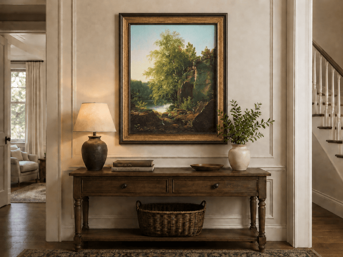 Framed hand-painted sunlit lakeside oil painting with rocks and leafy birch trees displayed above an entryway console table.